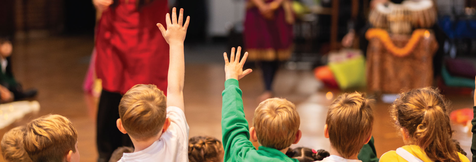 Five children watch a performer leading a workshop; two boys raise their hands.