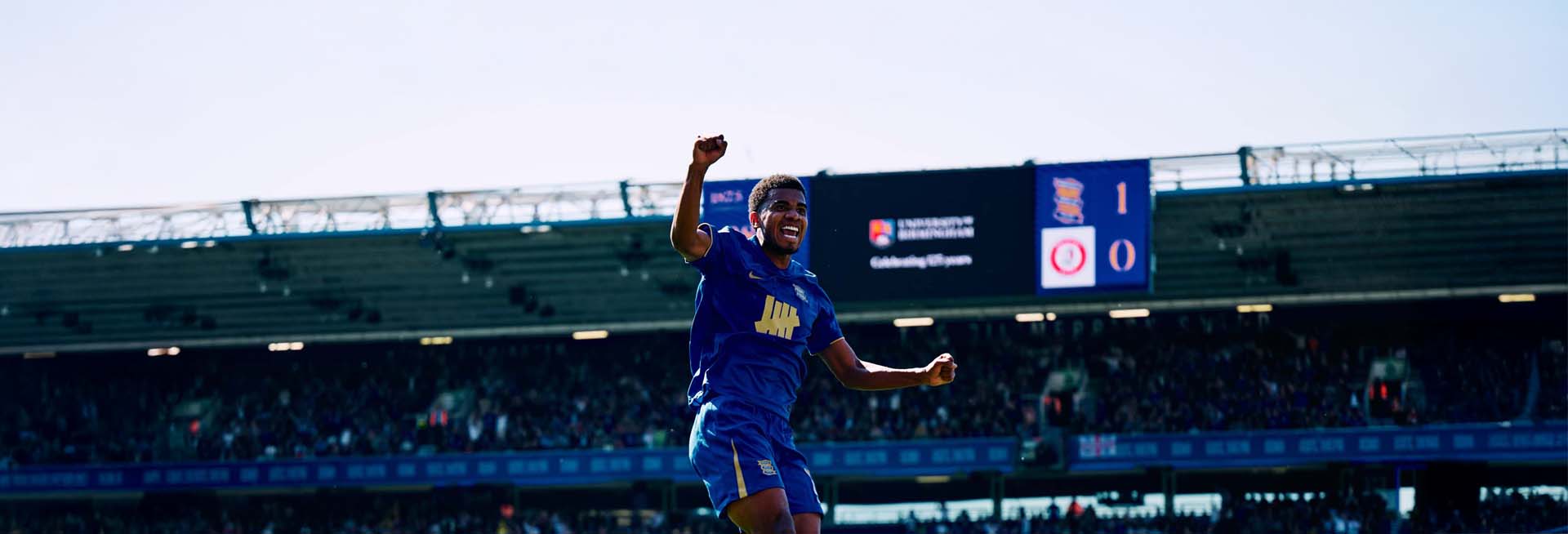 Birmingham City player Jhon Solís celebrating a goal, with University of Birmingham branding visible on the scoreboard