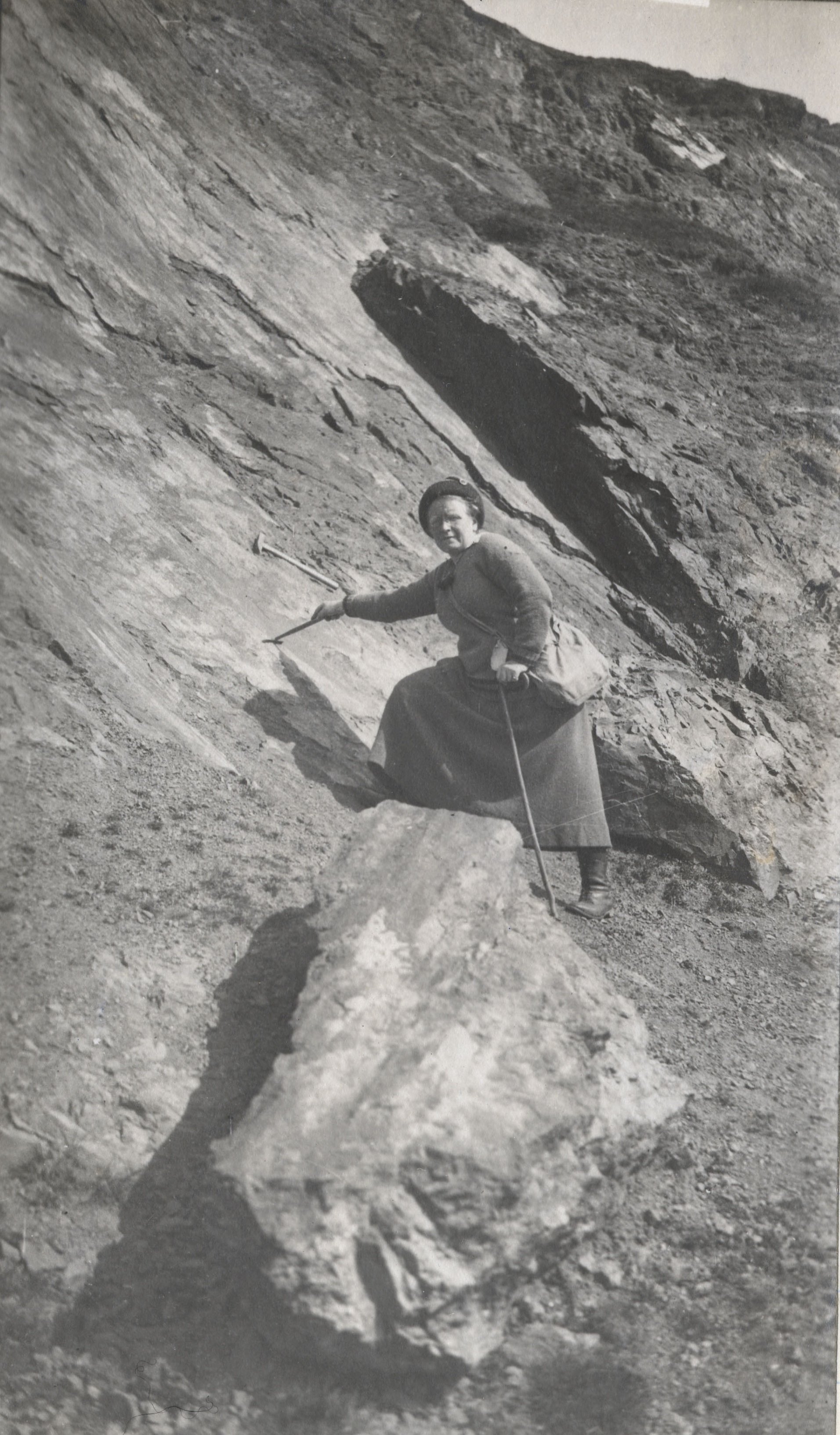 Gertrude climbs a slope in a dress, with a geological tools in hand.