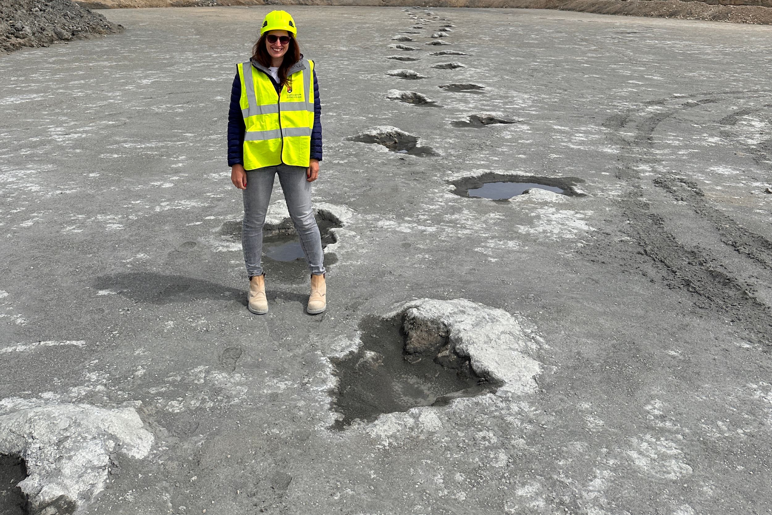 Kirsty Edgar stands in a hi-vis jacket beside a dinosaur fossil footprint trackway.