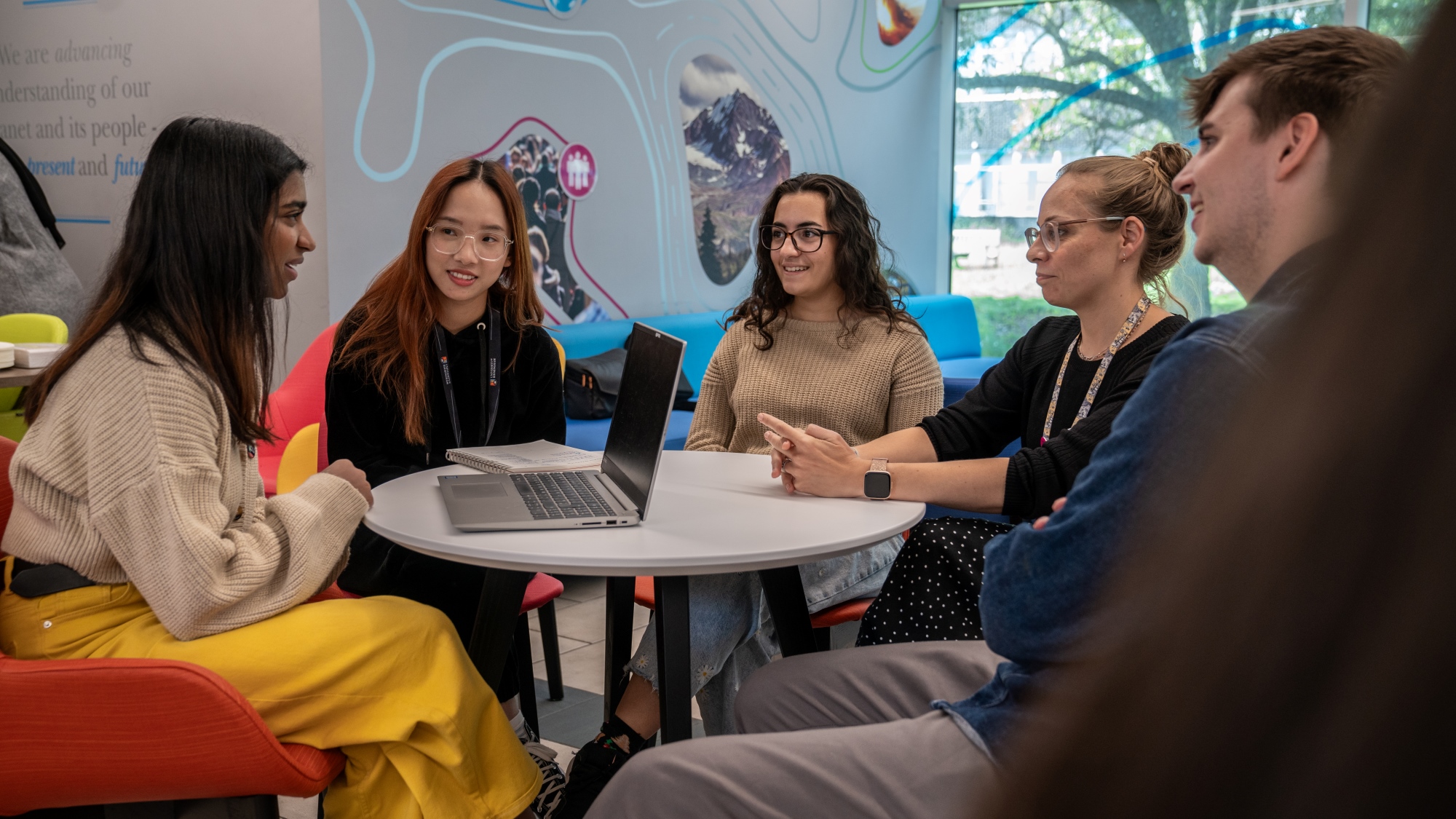 Five people in discussion around a table with laptop, near windows and colorful wall art.