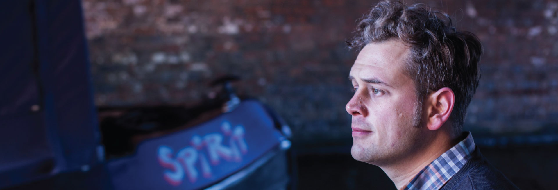 Author Luke Kennard wears a blue and white checked collar under a navy jumper and looks out to the left of frame.