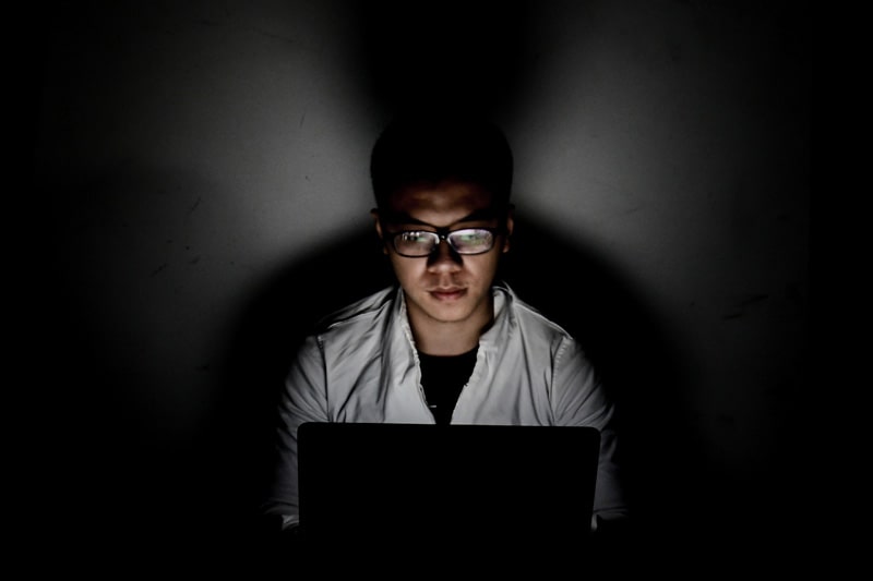 A man looking at a laptop in a darkened room with the bright screen illuminating his face
