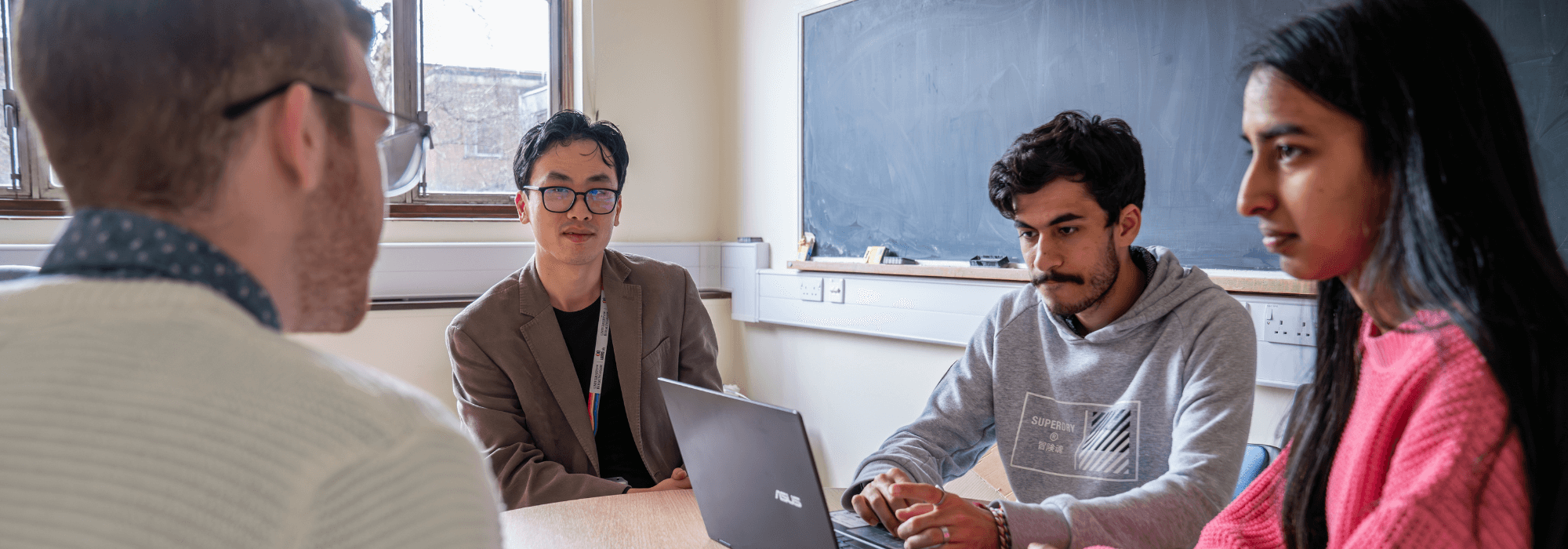 three students and an academic sitting around a small table with a laptop and blackboard in the background