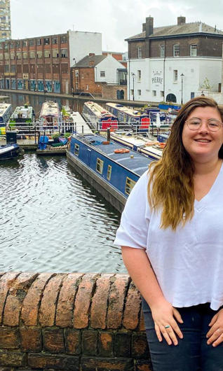 Lucy wears a white t-shirt and stands in front of canal boats.