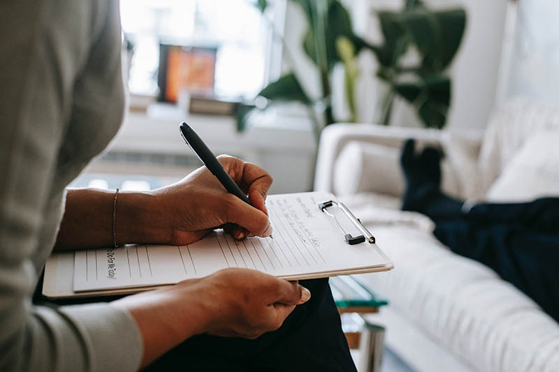 Woman writing on a clipboard next to person lying on a bed