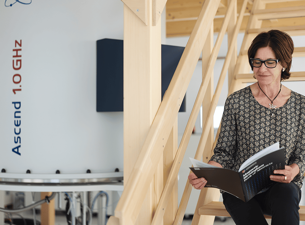 HWB-NMR Director Teresa Carlomagno sitting on wooden steps next to a 1.0 GHz spectrometer reading a University brochure.