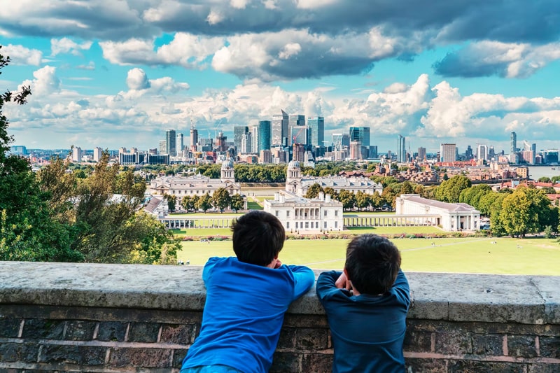 Two boys leaning on a wall looking out at the London skyline in the distance, defined against a blue, cloudy sky.