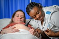 Midwife listening to a pregnant woman's stomach with a Pinard horn and checking her fob watch