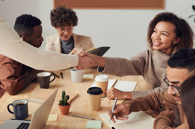 Two people shaking hands across a table in a group of people at a meeting