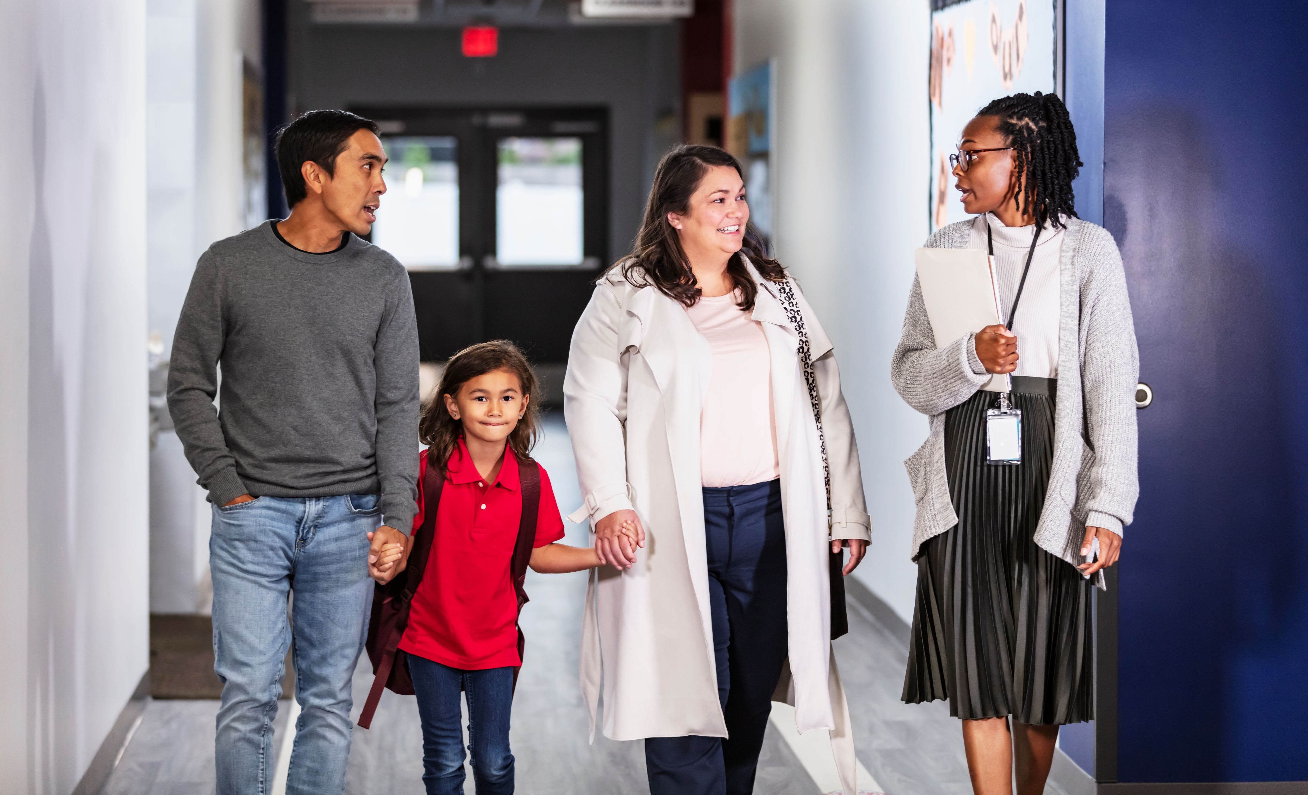 Parents holding their child's hands walking down a school corridor with a teacher