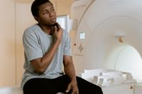 Young black man wearing a grey t-shirt and black trousers sitting on a bench of an fMRI machine waiting to speak to a doctor