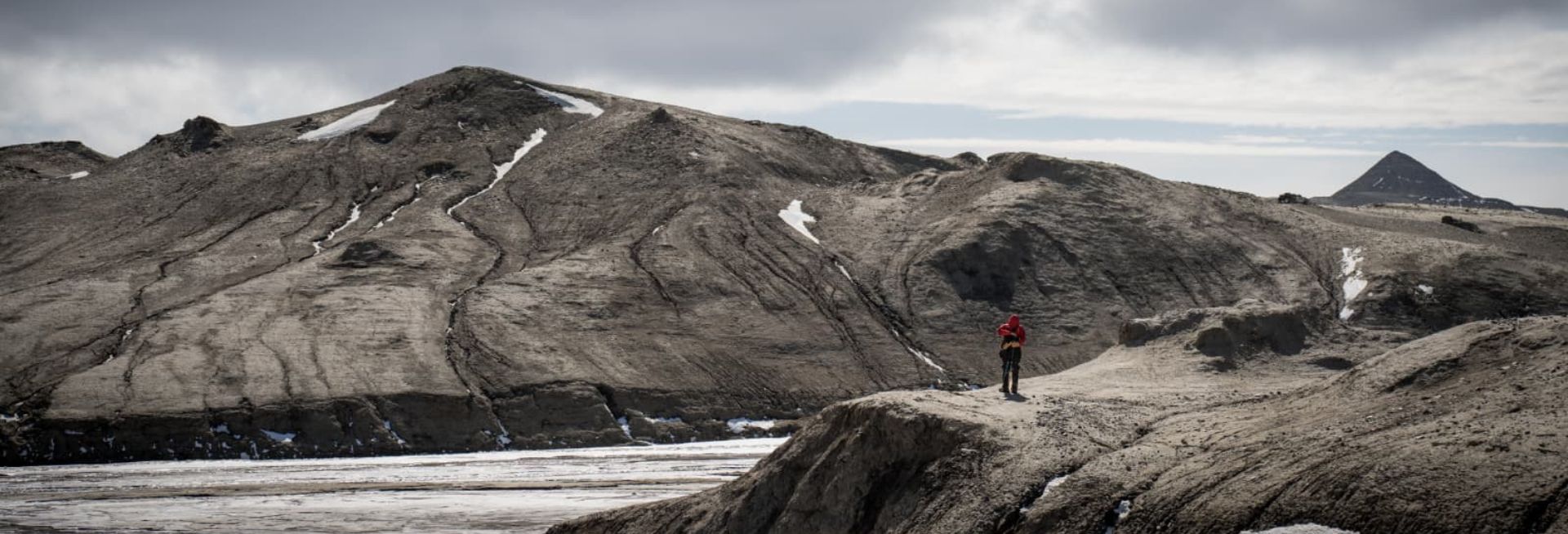 A photograph of a grey, cloudy mountainous landscape and a man in a red jacket and black trousers standing on a cliff in the foreground.