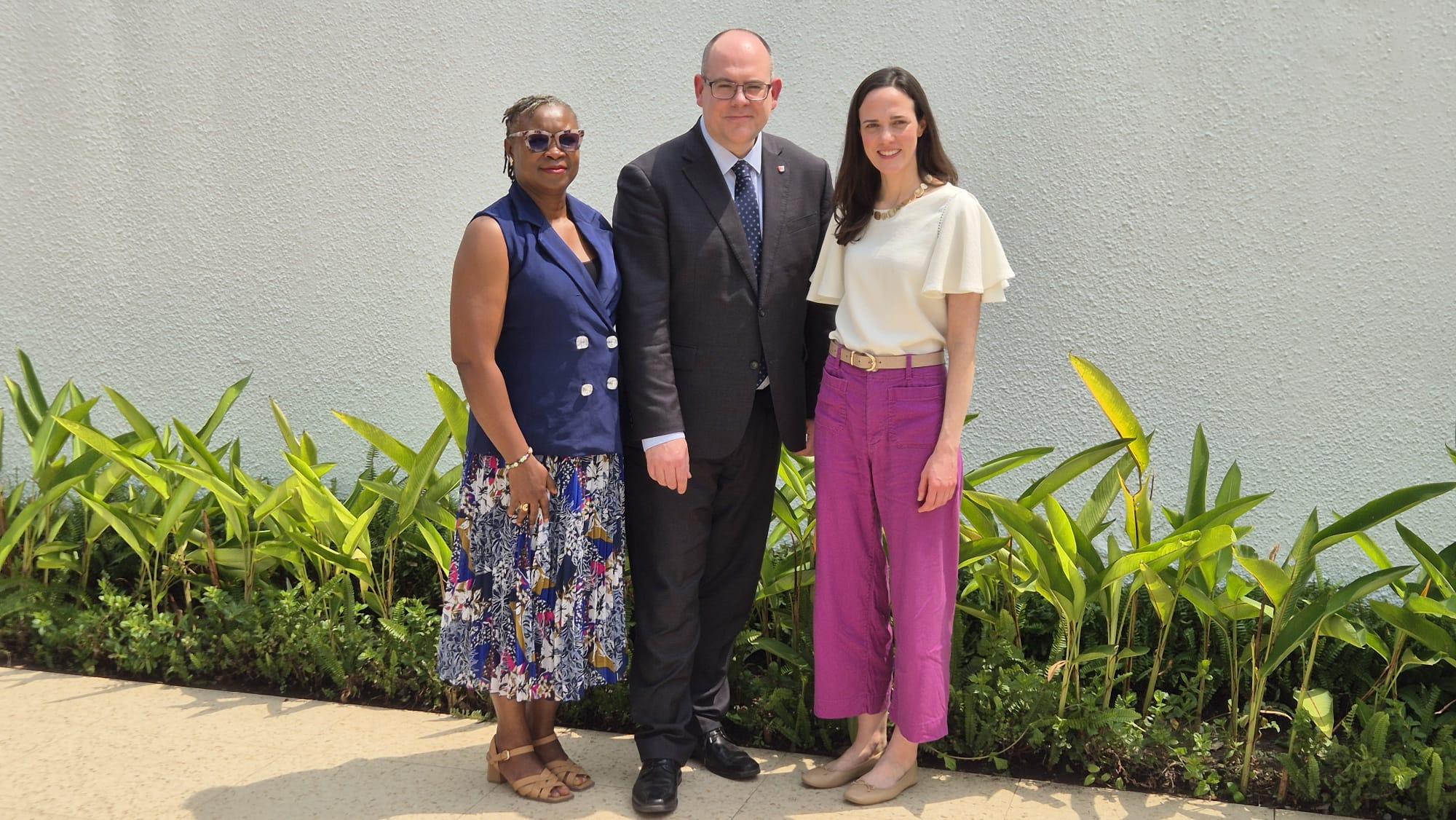 Three people standing against a white wall with plants.