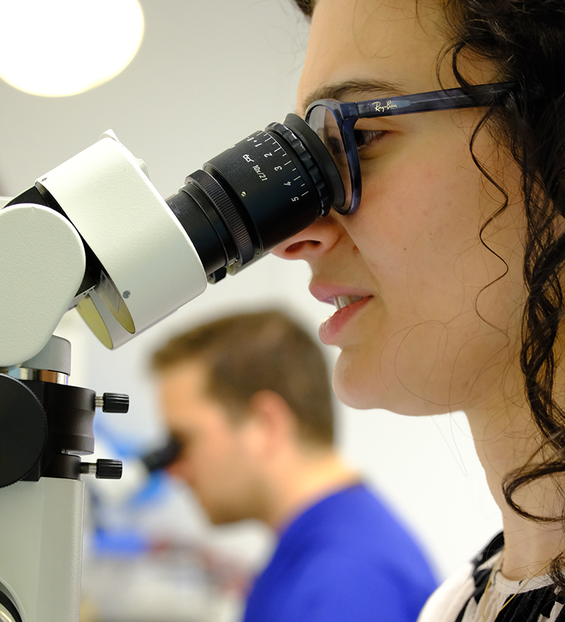 Close-up side view of a woman looking into a microscope with a male in the background also using a microscope.