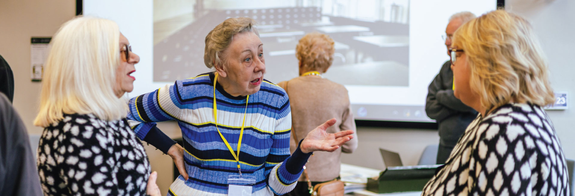 A white woman in a blue striped top and yellow lanyard speaks to two other white women.