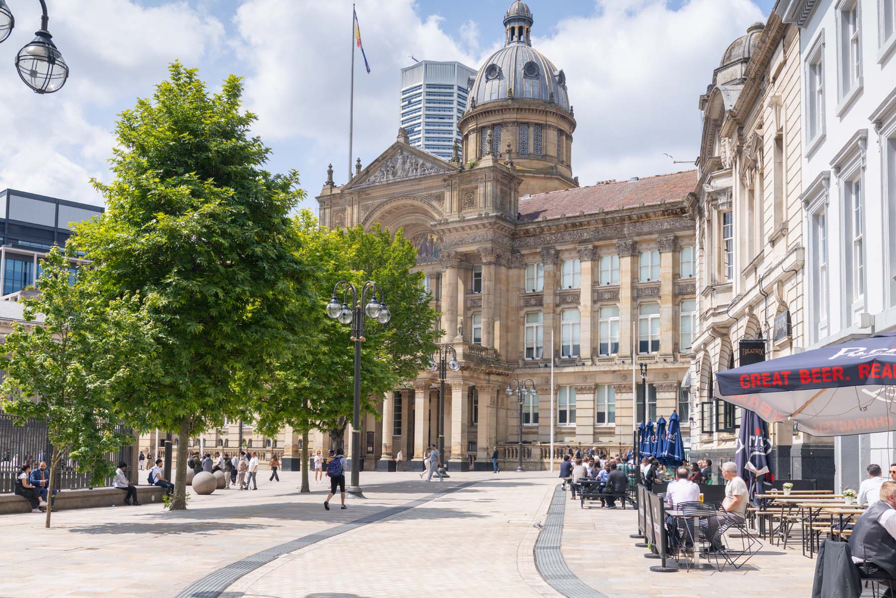 People sat outside on tables in front of greenery and sandstone building