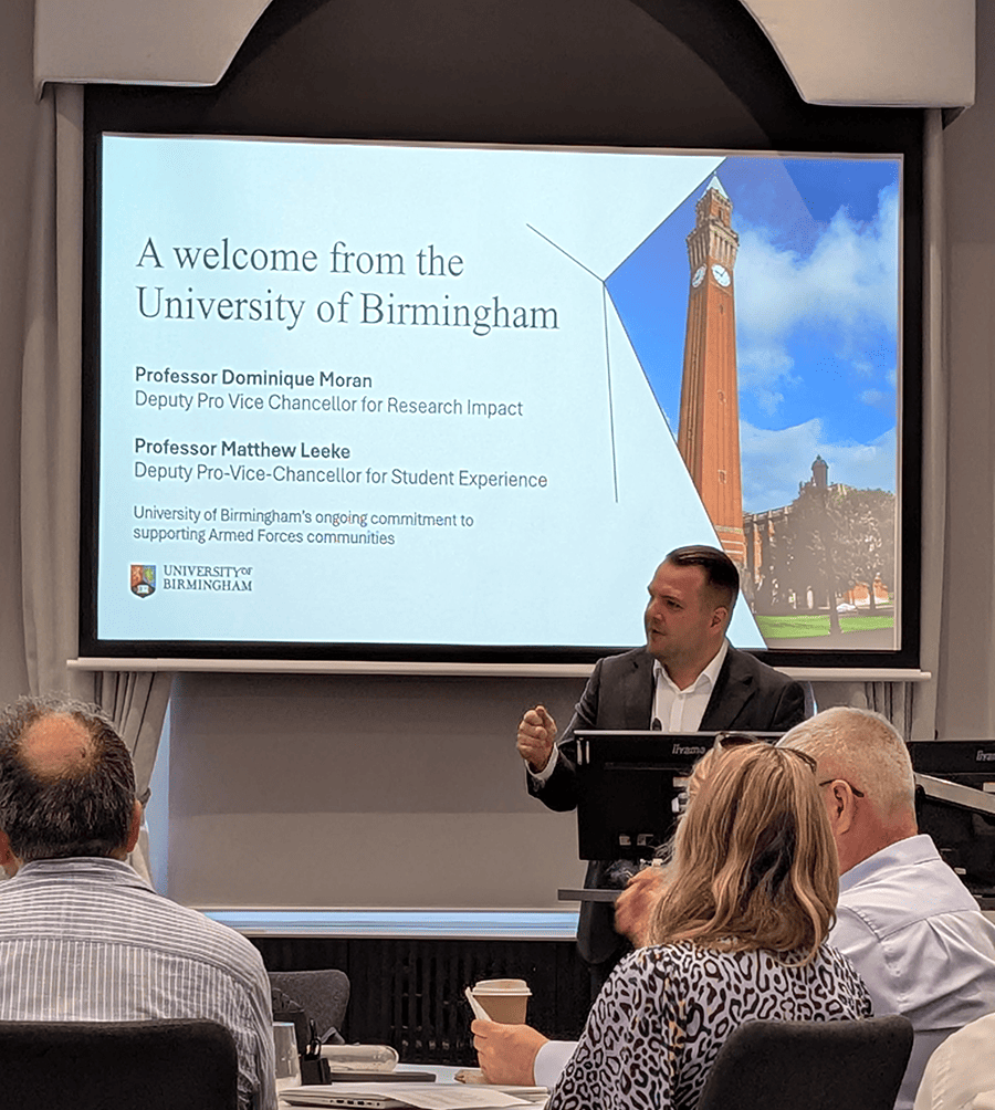 Professor Matt Leeke giving a presentation to a seated audience with PowerPoint slides behind him.