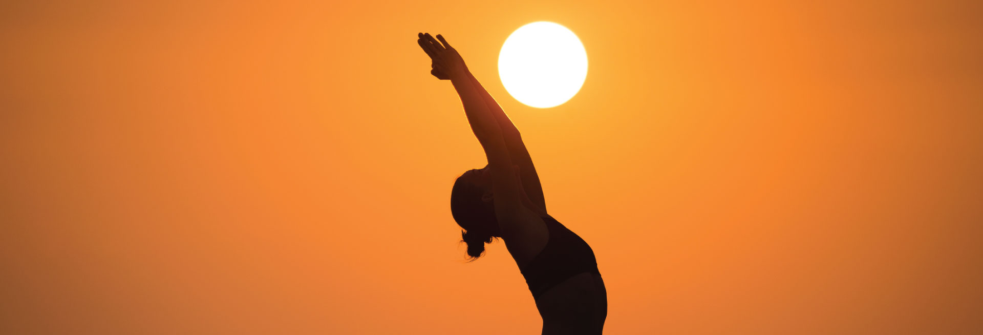 A woman doing a warrior yoga pose is silhouetted against a sunset.