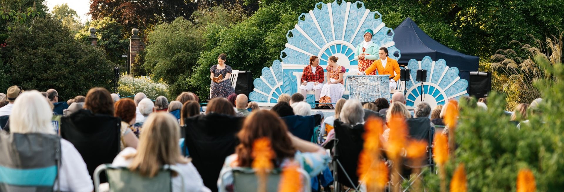 A blue stage set against a lush green backdrop, with actors in period costume. 