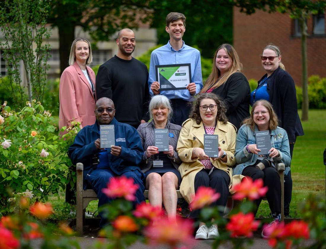 A group sit on a bench with a row of people behind them, all holding awards and smiling at the camera.