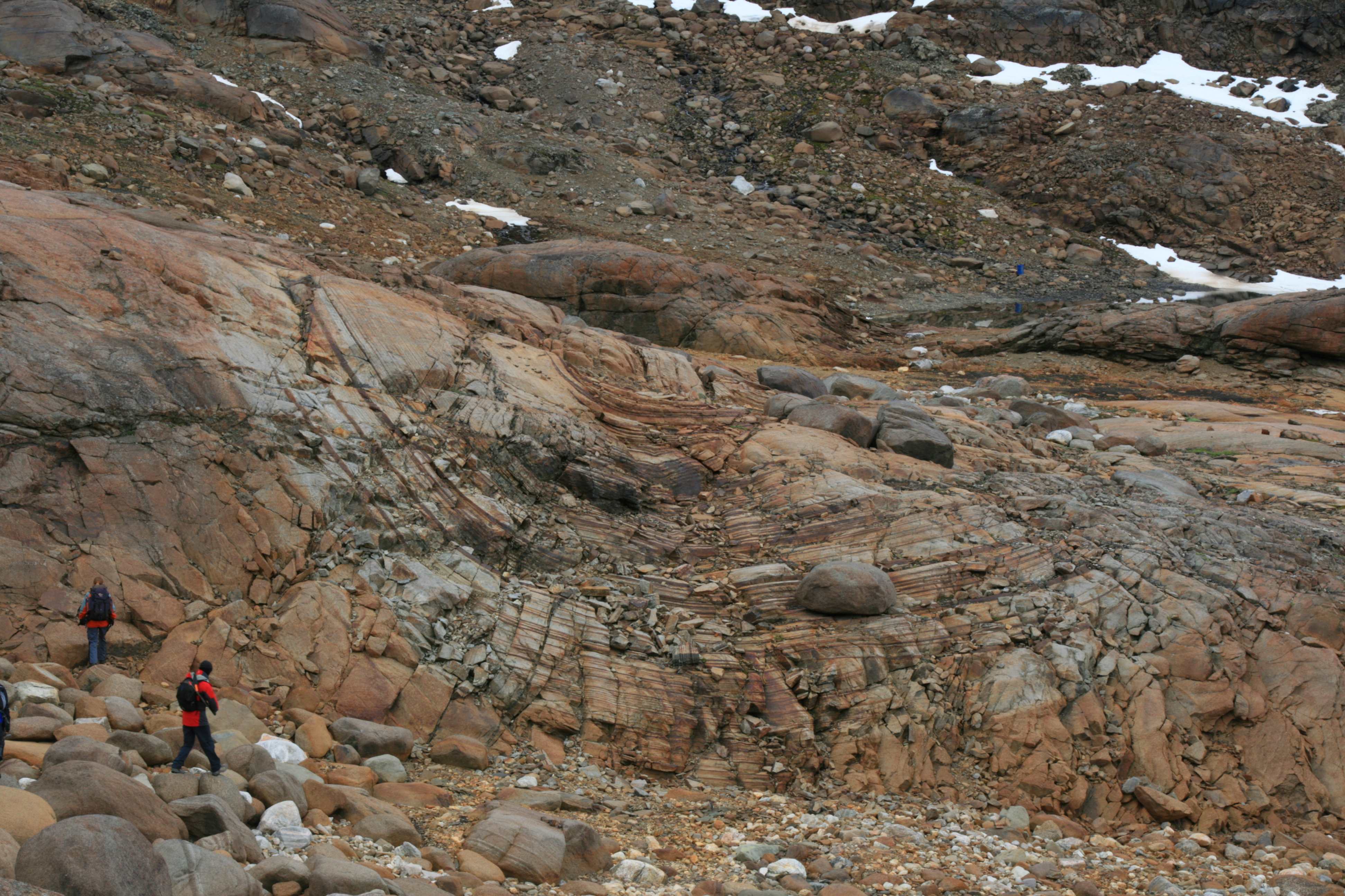A landscape of deformed rock beds and rocky outcrops. In the foreground are some people doing fieldwork.