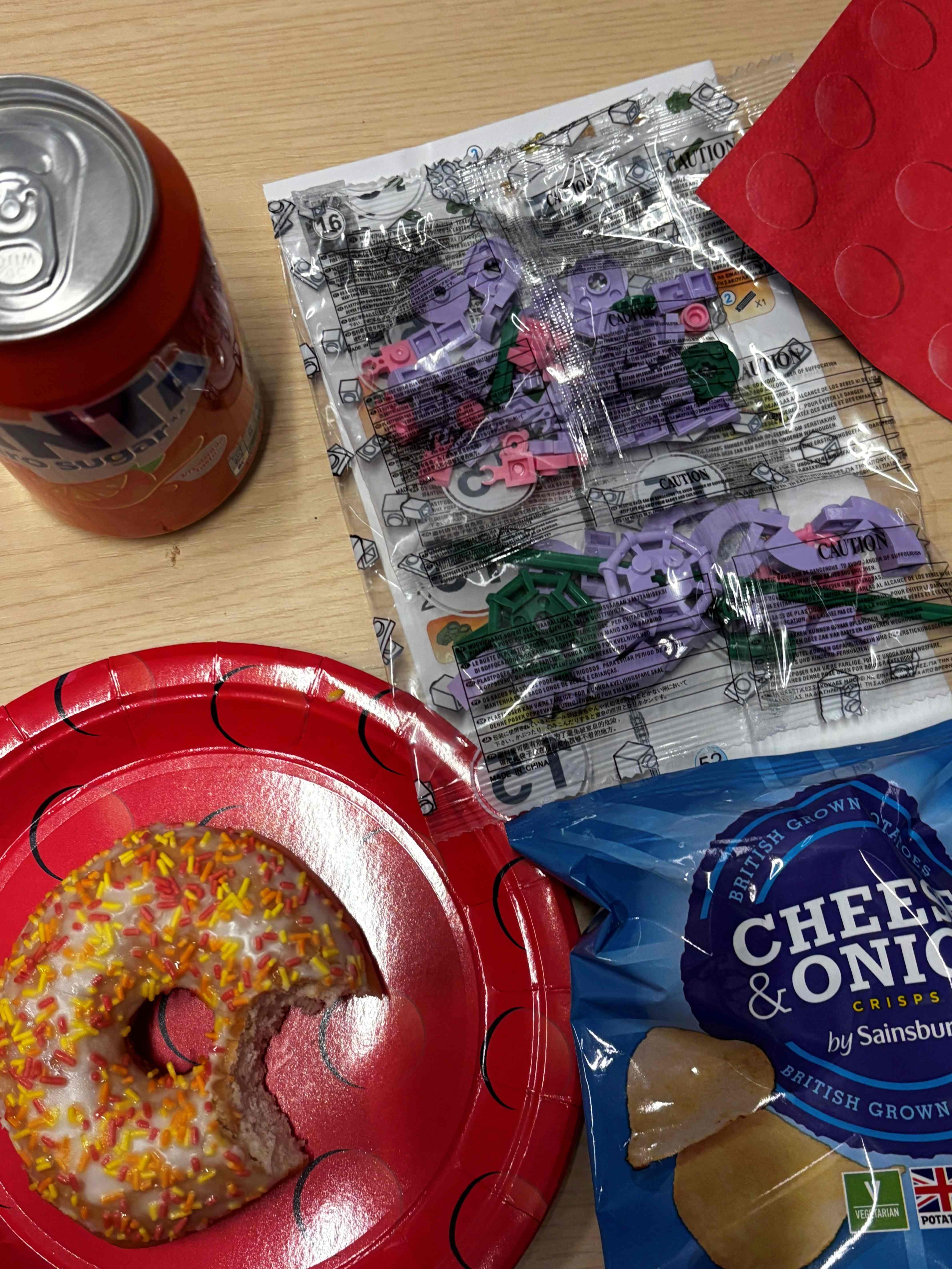 A table with a donut with sprinkles, a packet of cheese and onion crisps and a can of fanta