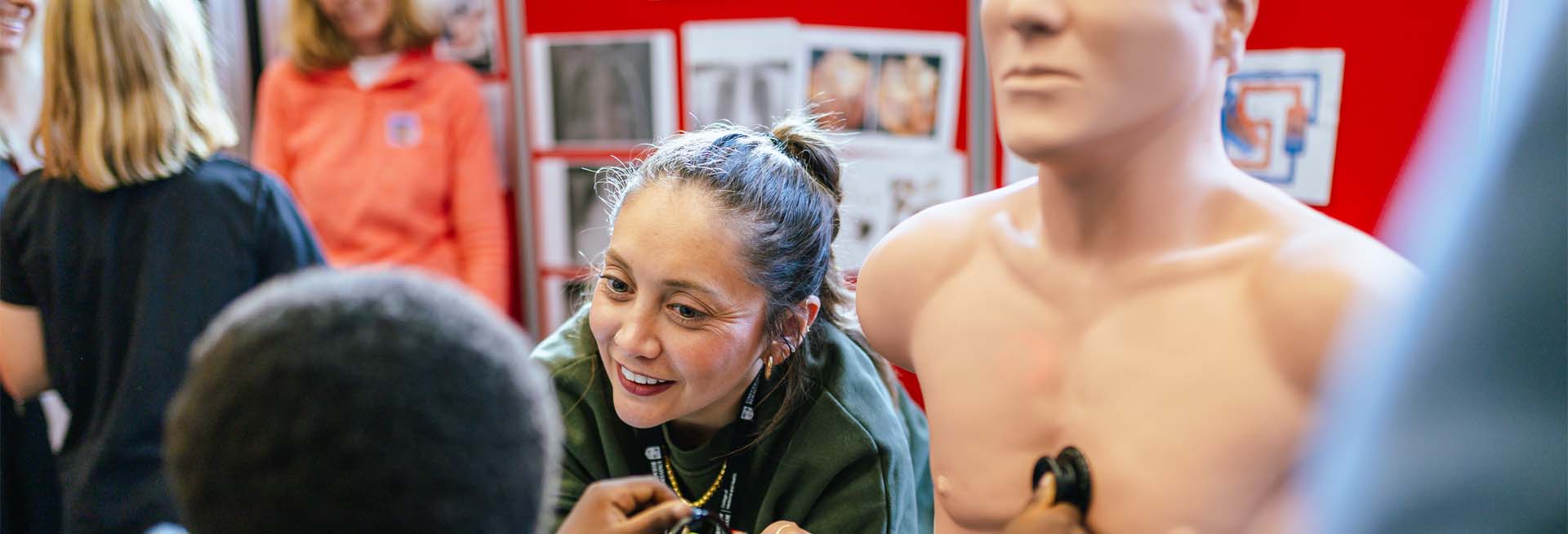 Child using a stethoscope on a dummy model to practice hearing a heartbeat at a public event.