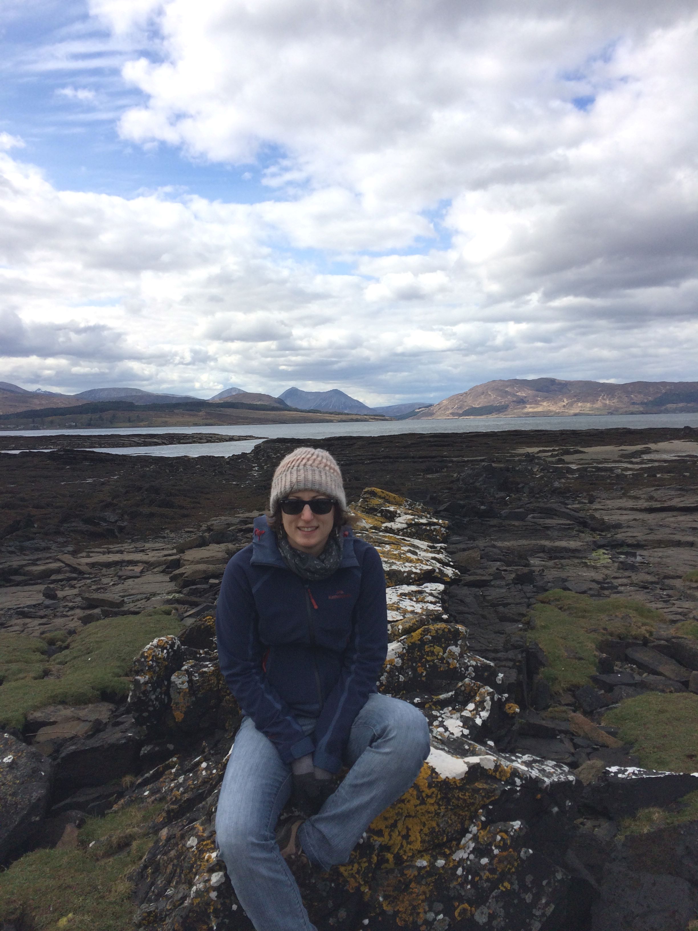A woman wearing sunglasses, a white hat, blue jacket and jeans sits on a rock. Behind her is a mountainous landscape with a lake.